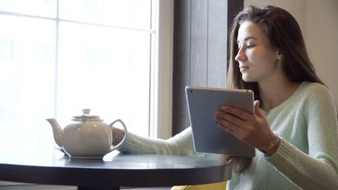 Girl sits at a table and drinking tea with a tablet pc in hands Stock Footage 70923095