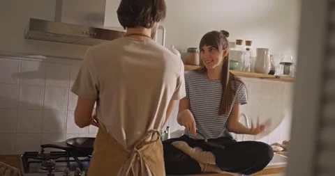 Girl sits on the table and talks to her boyfriend who is preparing breakfast in Stock Footage 318079420