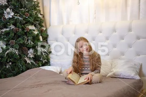 Girl sitting on the bed with a book by the christmas tree Stock Photos