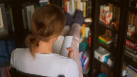 Girl Sitting by Bookcase Flips Through Softback Book Stock Footage