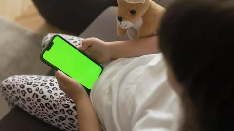 The girl is sitting on the couch and using a phone. The phone is in focus, with Stock-Footage 244477944
