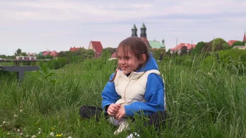 Girl sitting on the grass with a joyful expression, city in the background Stock Footage 309009751