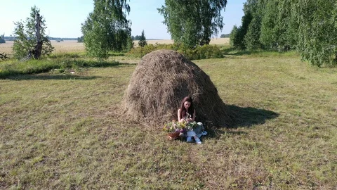 Girl sitting by a haystack on a hot summer day makes a wreath of flowers Stock Footage 128350509