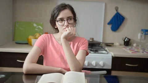 Girl sitting thinking at a table in the kitchen 스톡 동영상 129880843