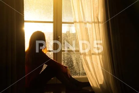 Girl sitting on a windowsill and looking at the evening city Stock Photos
