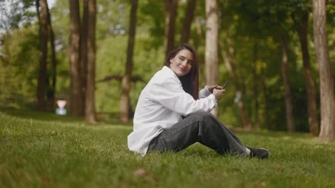 Girl student sitting on grass, using mobile phone, waving hello to friends Stock Footage 235930832