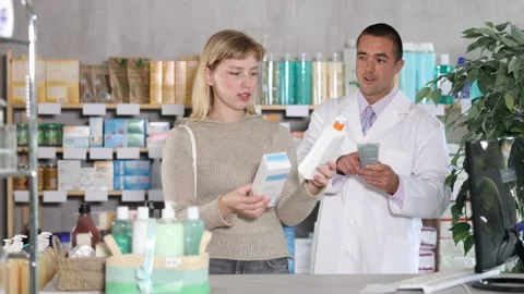 Girl studies composition of body lotion, man pharmacist in background Stock Footage 321720637