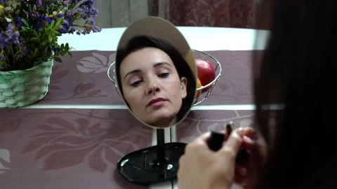 The girl at the table looks into her reflection in a small mirror and paints her Stock Footage 144026552