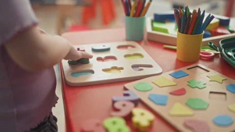 Girl toddler with small hand places a wooden puzzle shape on a red table pu.. Stock Footage 339251053