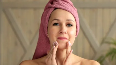 A girl with a towel on her head and bare shoulders puts cream on her face and Stock Footage