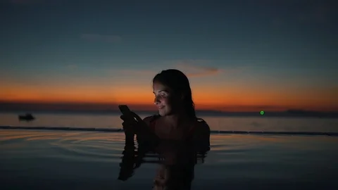 Girl types message on smartphone in the pool at sunset Stock Footage 110187436