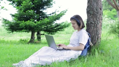 The girl is typing on a laptop while sitting near a tree in the park Stock Footage 159386763