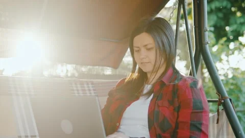 Girl typing on a laptop while sitting on a swing in the garden in the backyard. Video stock 244647802