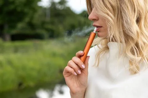 Girl using a vape device while standing near a small body of water in a gre.. Stock Photos