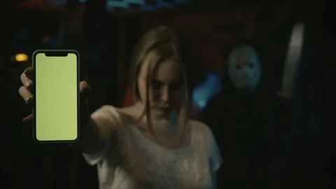 Girl victim standing in the warehouse and holding a smartphone. Man in hockey Stock Footage 248058341