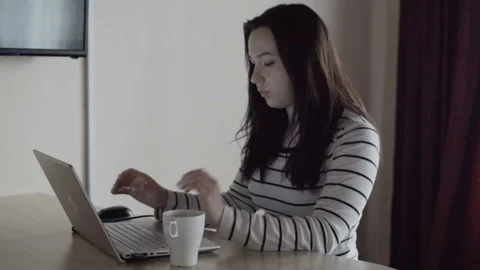 The girl in a white jacket works at the computer. Drinking hot morning coffee. Video stock 96114483
