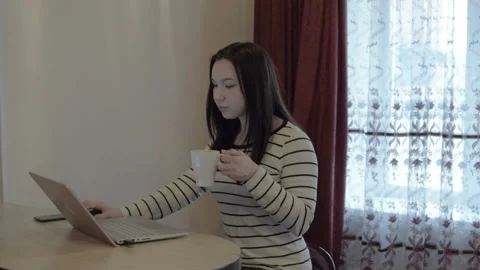 The girl in a white jacket works at the computer. Drinking hot morning coffee. 스톡 동영상 96114548
