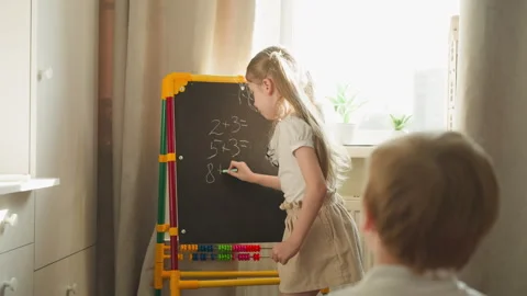 Girl writes math problems for brother playing school at home Stock Footage 248743830