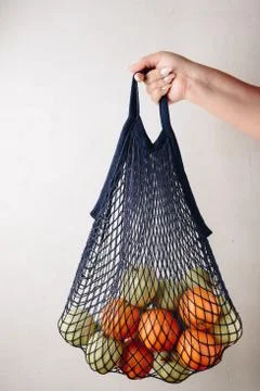 The girl's hand holds a blue string bag with green apples and oranges on a gr 스톡 사진