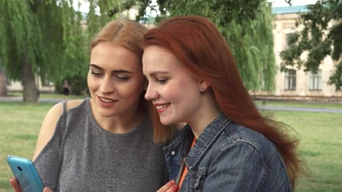 Girls joyfully sharing a smartphone while chatting in the park Stock Footage 311980827