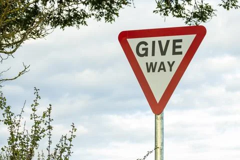 Give way warning red triangle sign beside hedge and trees on country road Stock Photos