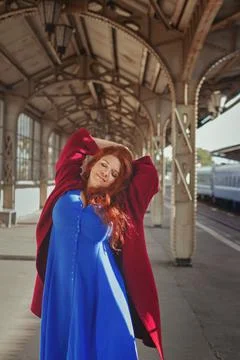 Glad young smiling red-haired European woman in red coat and blue dress at train Stock Photos