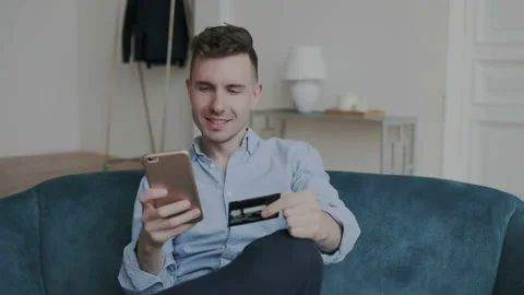 Glamorous and charming young man buying online uses a phone and a credit card on Stock Footage 107788807