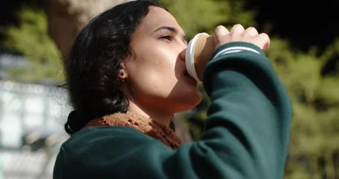 Glancing right, woman in varsity jacket raising coffee cup to sip in park, copy Stock Footage 317809185