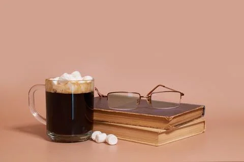 A glass cup of coffee with marshmallows, stack of old books and glasses Stock Photos