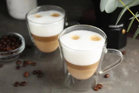 Glass cups of delicious latte macchiato and scattered coffee beans on grey ta Stock Photos
