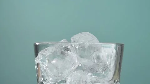 Glass filled with ice cubes slowly rotates on turquoise background, evoking a Stock Footage 306165887