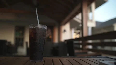 Glass of iced beverage with straw, placed on wooden table in outdoor setting Video stock 295168512