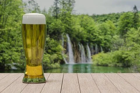 Glass of light beer on table with view of forest with lake and waterfall Stock Photos