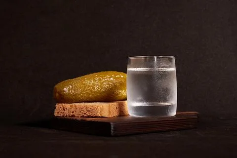 A glass of vodka, rye bread and a pickle on a wooden stand Stock Photos
