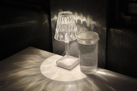 A glass of water sits on a table next to a lamp Stock Photos