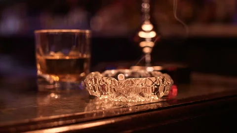 Glass of whiskey and burning cigarettes resting on an ashtray Stock Footage 87102003