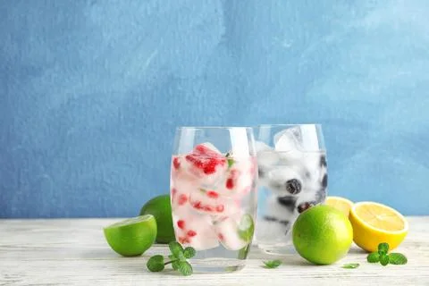 Glasses of drinks with fruit ice cubes on table against color background Stock Photos