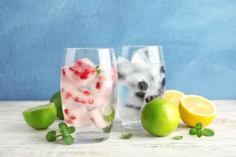 Glasses of drinks with fruit ice cubes on table against color background Stock Photos