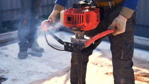Gloved construction worker operating red earth auger outdoors, drilling hole Stock Photos