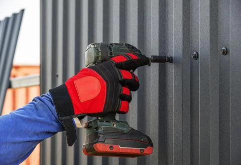 A gloved construction worker uses a drill driver to drive a screw into a dark Foto stock