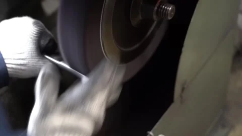 Gloved craftsman grinds a slender steel rod against a spinning abrasive wheel Stock Footage 346297828