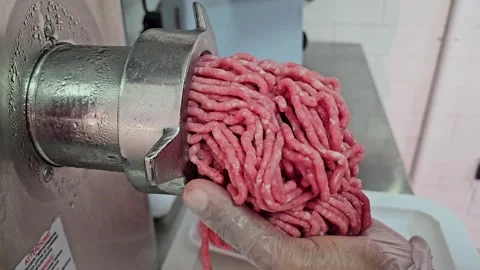 A gloved hand catches freshly ground pink meat emerging from a metal grinder Stock Footage 311576342