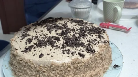 Gloved hand decorating beige frosted cake with crumb topping on glass plate in Video stock 343516081