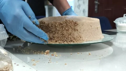 Gloved hand decorating beige frosted cake with crumb topping on glass plate in Video stock 343516236