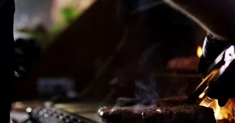 Gloved Hand Grilling Meat over Barbecue Flames with Tongs, Dark Fire Close Up Stock Footage 340321700