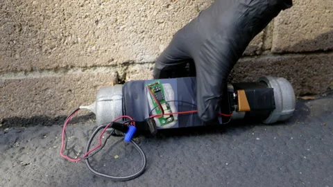 GLOVED HAND PLACING A PIPE BOMB.  4K SLIDER SHOT Stock Footage 163563162
