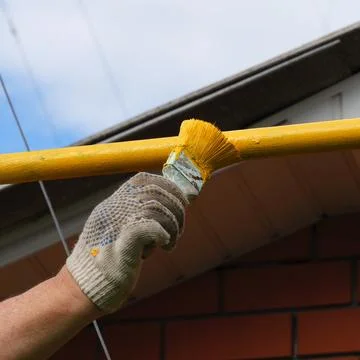 A gloved hand using a brush to apply yellow paint to a metal pipe outdoors. Stock-Fotos
