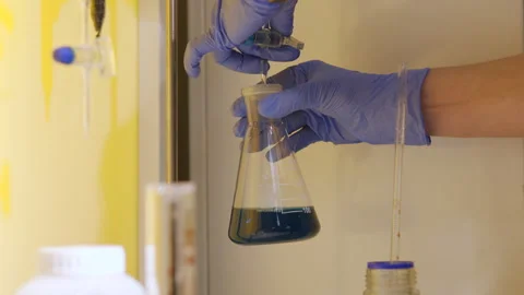 Gloved hands adding reagent drop into Erlenmeyer flask with blue solution inside Stock Footage 348261950
