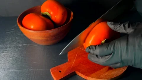 Gloved hands cutting ripe persimmon with sharp knife on wooden cutting board Stock Footage 325956574