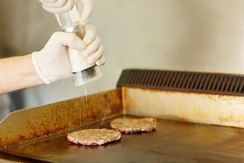 Gloved hands grinding salt onto two frying hamburger patties Stock Photos